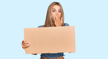 Young blonde woman holding cardboard empty banner covering mouth with hand, shocked and afraid for...