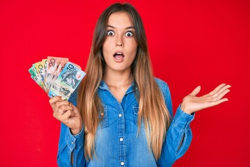 Beautiful caucasian woman holding australian dollars scared and amazed with open mouth for surprise, disbelief face