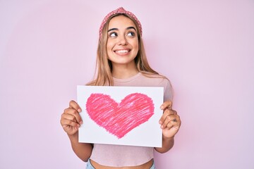 Young beautiful blonde woman holding heart draw smiling looking to the side and staring away thinking.