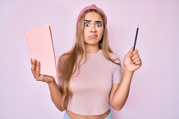 Young beautiful blonde woman holding book and pencil skeptic and nervous, frowning upset because of...