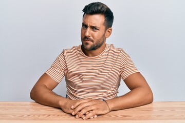 Handsome hispanic man wearing casual clothes sitting on the table skeptic and nervous, frowning...