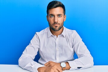 Handsome hispanic man wearing business clothes sitting on the table relaxed with serious expression...