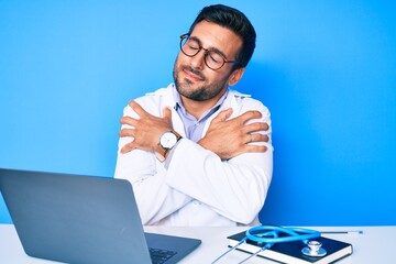 Young hispanic man wearing doctor uniform working at the clinic hugging oneself happy and positive,...