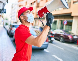 Young delivery man wearing uniform and coronavirus protection medical mask. Screaming using megaphone at town street.