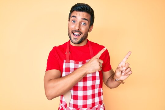 Young hispanic man wearing professional baker apron smiling and looking at the camera pointing with two hands and fingers to the side.