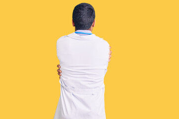 Young hispanic man wearing doctor uniform and stethoscope hugging oneself happy and positive from...