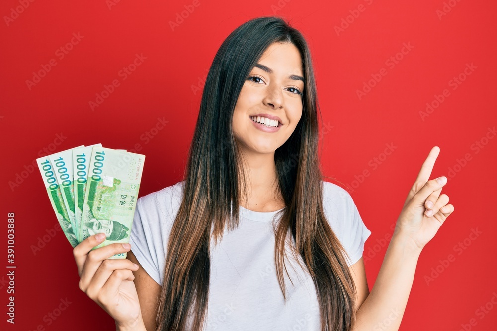 Sticker Young brunette woman holding 1000 south korean won banknotes smiling happy pointing with hand and finger to the side