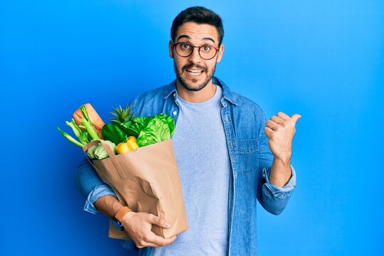 Young hispanic man holding paper bag with bread and groceries pointing thumb up to the side smiling happy with open mouth