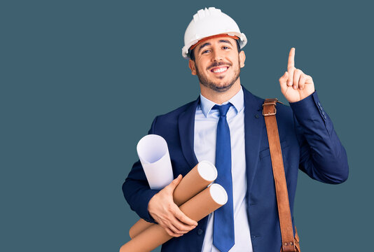 Young hispanic man wearing architect hardhat and leather bag holding blueprints surprised with an idea or question pointing finger with happy face, number one