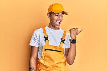 Young handsome african american man wearing handyman uniform over yellow background smiling with happy face looking and pointing to the side with thumb up.
