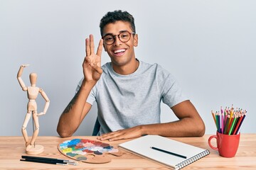 Young handsome african american man painter sitting palette and art manikin showing and pointing up with fingers number three while smiling confident and happy.