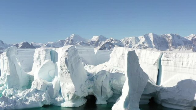 4K Aerial Landscape of snowy mountains and icy shores in Antarctica. Beautiful blue iceberg with mirror reflection floats in open ocean. Glaciers of a harsh continent.