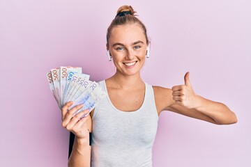 Beautiful blonde sport woman holding 20 swedish krona banknotes smiling happy and positive, thumb up doing excellent and approval sign