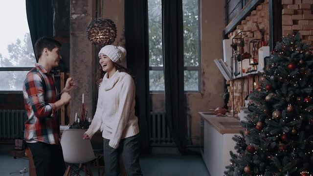 Lively young couple wearing winter sweaters listening to funky music dancing having fun in a brightly decorated living room decorated with bright Christmas tree lights. Happy winter holidays and hobby