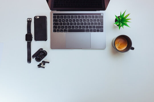 Top view of gray laptop with coffee cup, small plant, smartphone, wifi earphones and smartwatch on white background