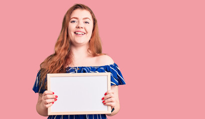 Fototapeta na wymiar Young beautiful redhead woman holding empty white chalkboard looking positive and happy standing and smiling with a confident smile showing teeth