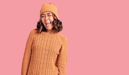 Young beautiful mixed race woman wearing wool sweater and winter hat winking looking at the camera with sexy expression, cheerful and happy face.