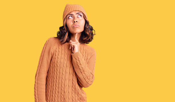Young beautiful mixed race woman wearing wool sweater and winter hat thinking concentrated about doubt with finger on chin and looking up wondering