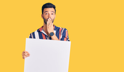 Young handsome hispanic man holding cardboard banner with blank space covering mouth with hand, shocked and afraid for mistake. surprised expression