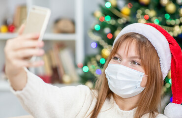 Young woman wearing Christmas hat, chatting with smartphone at home, with Christmas tree on the background. Coronavirus and Christmas concept