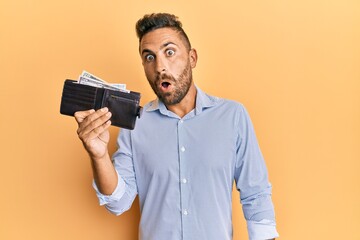 Handsome man with beard holding wallet with dollars scared and amazed with open mouth for surprise, disbelief face