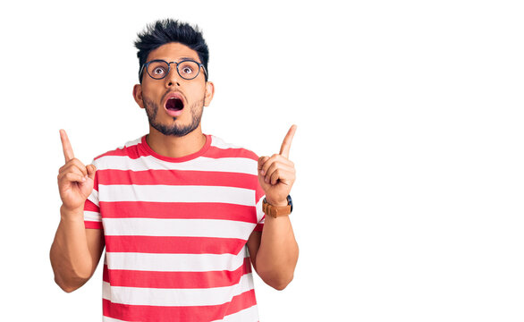 Handsome latin american young man wearing casual clothes and glasses amazed and surprised looking up and pointing with fingers and raised arms.