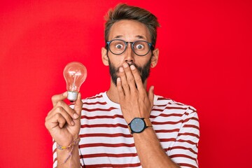 Handsome caucasian man with beard holding lightbulb for inspiration and idea covering mouth with...