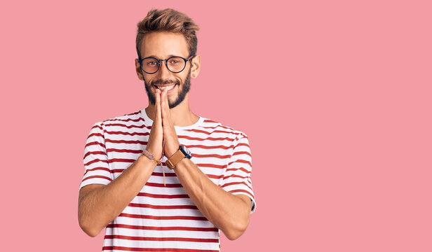 Handsome blond man with beard wearing casual clothes and glasses praying with hands together asking for forgiveness smiling confident.