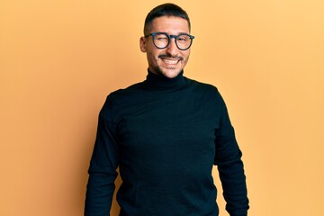 Handsome man with tattoos wearing turtleneck sweater and glasses winking looking at the camera with sexy expression, cheerful and happy face.