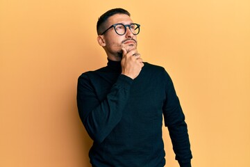 Handsome man with tattoos wearing turtleneck sweater and glasses with hand on chin thinking about question, pensive expression. smiling with thoughtful face. doubt concept.