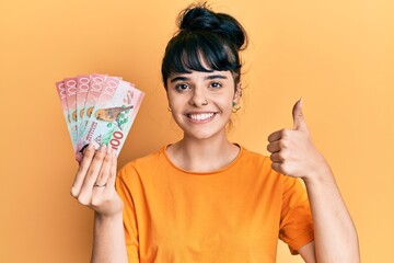 Young hispanic girl holding 100 new zealand dollars banknote smiling happy and positive, thumb up doing excellent and approval sign
