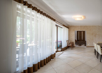 Modern brown interior of spacious apartment. Tulle curtain and panorama windows. Tile floor.