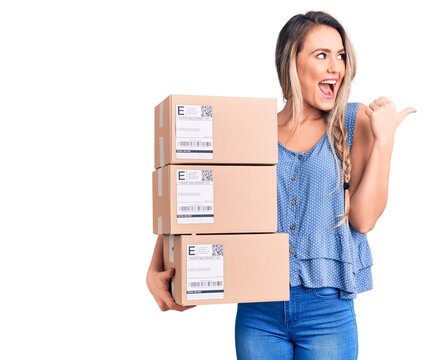 Young beautiful blonde woman holding delivery boxes pointing thumb up to the side smiling happy with open mouth