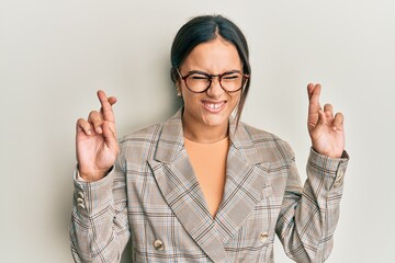 Young brunette woman wearing business jacket and glasses gesturing finger crossed smiling with hope and eyes closed. luck and superstitious concept.