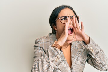 Young brunette woman wearing business jacket and glasses shouting angry out loud with hands over mouth