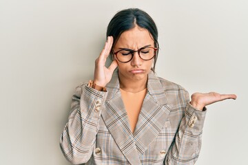 Young brunette woman wearing business jacket and glasses confused and annoyed with open palm showing copy space and pointing finger to forehead. think about it.
