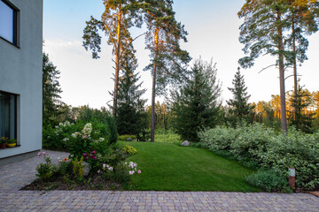 Modern exterior of private cottage near the pine forest. View of beautiful garden with flowers, bushes and trees.