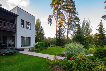 Modern exterior of private cottage near the pine forest. View of beautiful garden with flowers, bushes and trees.