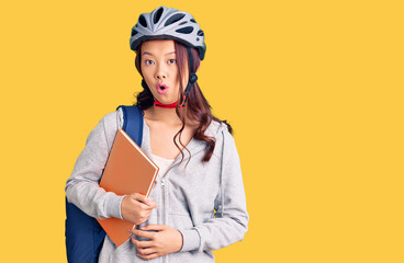 Young beautiful chinese girl wearing student backpack and bike helmet holding book scared and amazed with open mouth for surprise, disbelief face