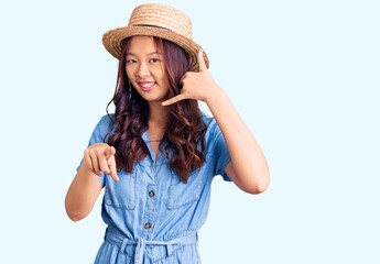 Young beautiful chinese girl wearing summer hat smiling doing talking on the telephone gesture and pointing to you. call me.