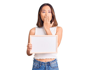 Young beautiful chinese girl holding empty white chalkboard covering mouth with hand, shocked and...