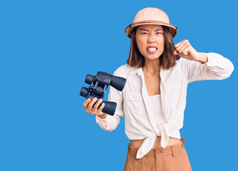 Young beautiful chinese girl wearing explorer hat holding binoculars annoyed and frustrated shouting with anger, yelling crazy with anger and hand raised