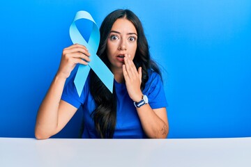 Beautiful brunette young woman holding blue ribbon covering mouth with hand, shocked and afraid for...