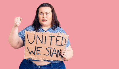 Young plus size woman holding united we stand banner annoyed and frustrated shouting with anger, yelling crazy with anger and hand raised