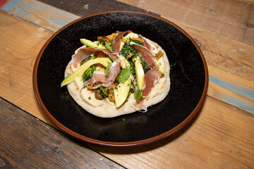 A delicious plate of a naan flatbread with Hummus, Mushrooms, Serrano Ham and Avocado on a wooden kitchen work top