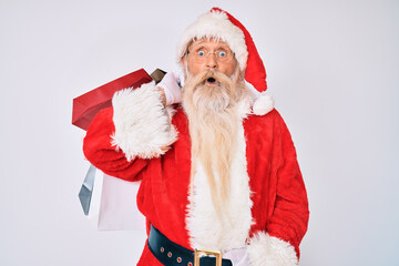 Old senior man with grey hair and long beard wearing santa claus costume holding shopping bags scared and amazed with open mouth for surprise, disbelief face