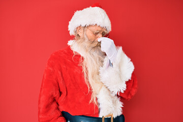 Old senior man with grey hair and long beard wearing traditional santa claus costume smelling something stinky and disgusting, intolerable smell, holding breath with fingers on nose. bad smell