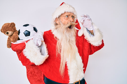 Old senior man with grey hair and long beard wearing santa claus costume holding mustache angry and mad screaming frustrated and furious, shouting with anger. rage and aggressive concept.