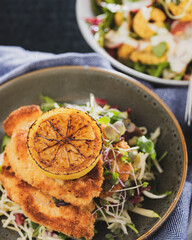A serving of Chicken Schnitzel in a bowl with fresh coleslaw, and a fried lemon wedge.
