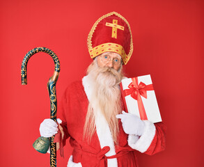 Old senior man with grey hair and long beard wearing saint nicholas costume holding present...